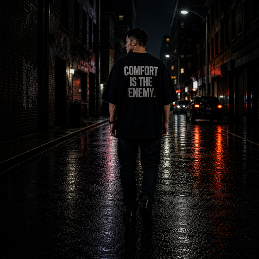Person wearing a black t-shirt with 'COMFORT IS THE ENEMY' text on a dark city street.