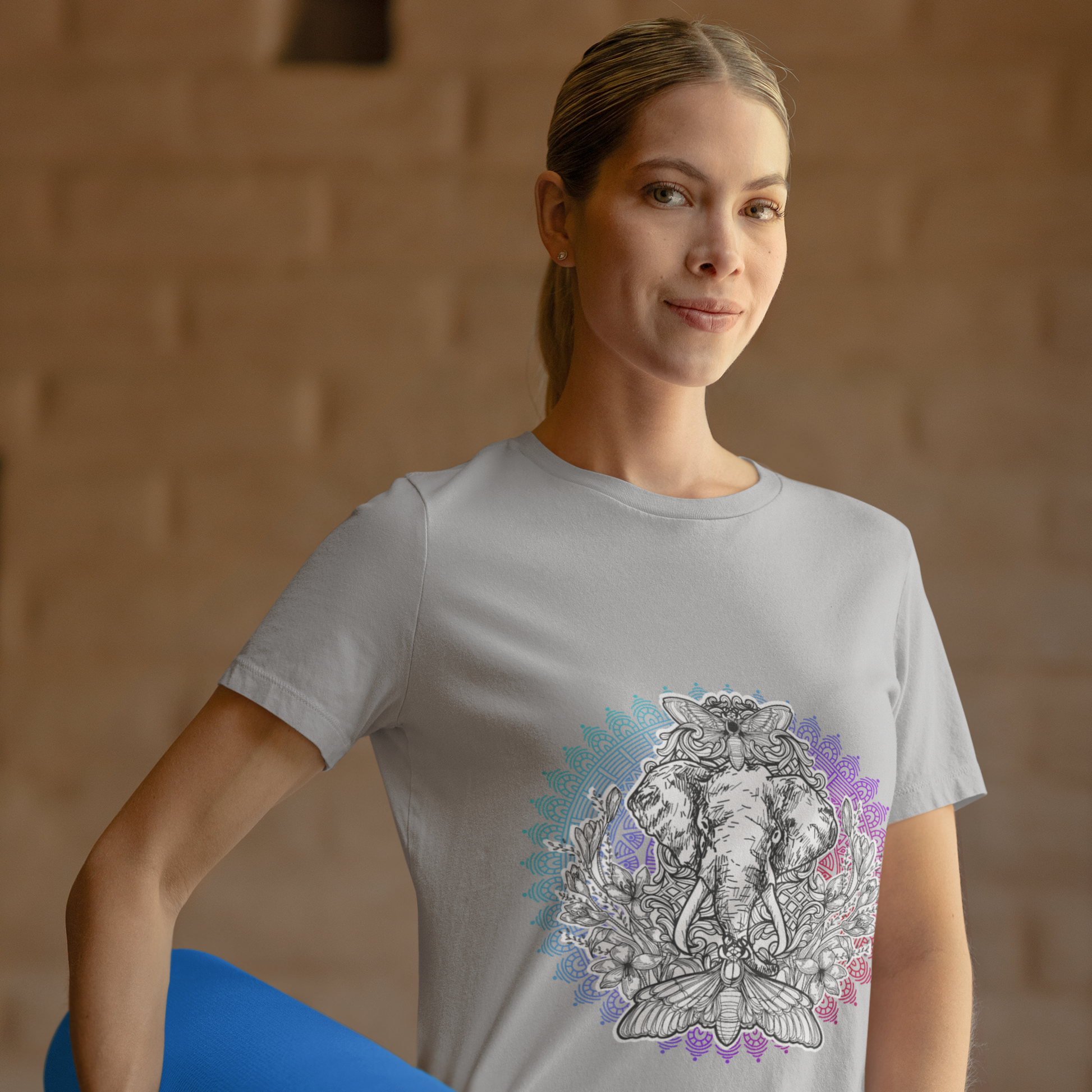 Woman wearing a t-shirt with a graphic design, holding a blue yoga mat against a brick wall.