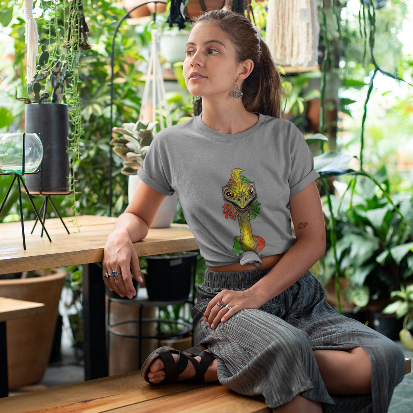 Woman sitting in a plant-filled room wearing a gray t-shirt with a colorful graphic design.