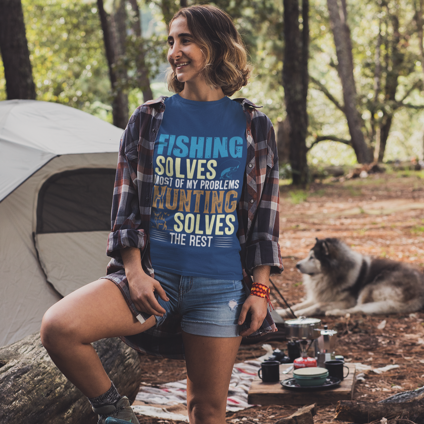 Woman in a blue t-shirt with outdoor text, sitting by a campsite with a dog and tent in the background.