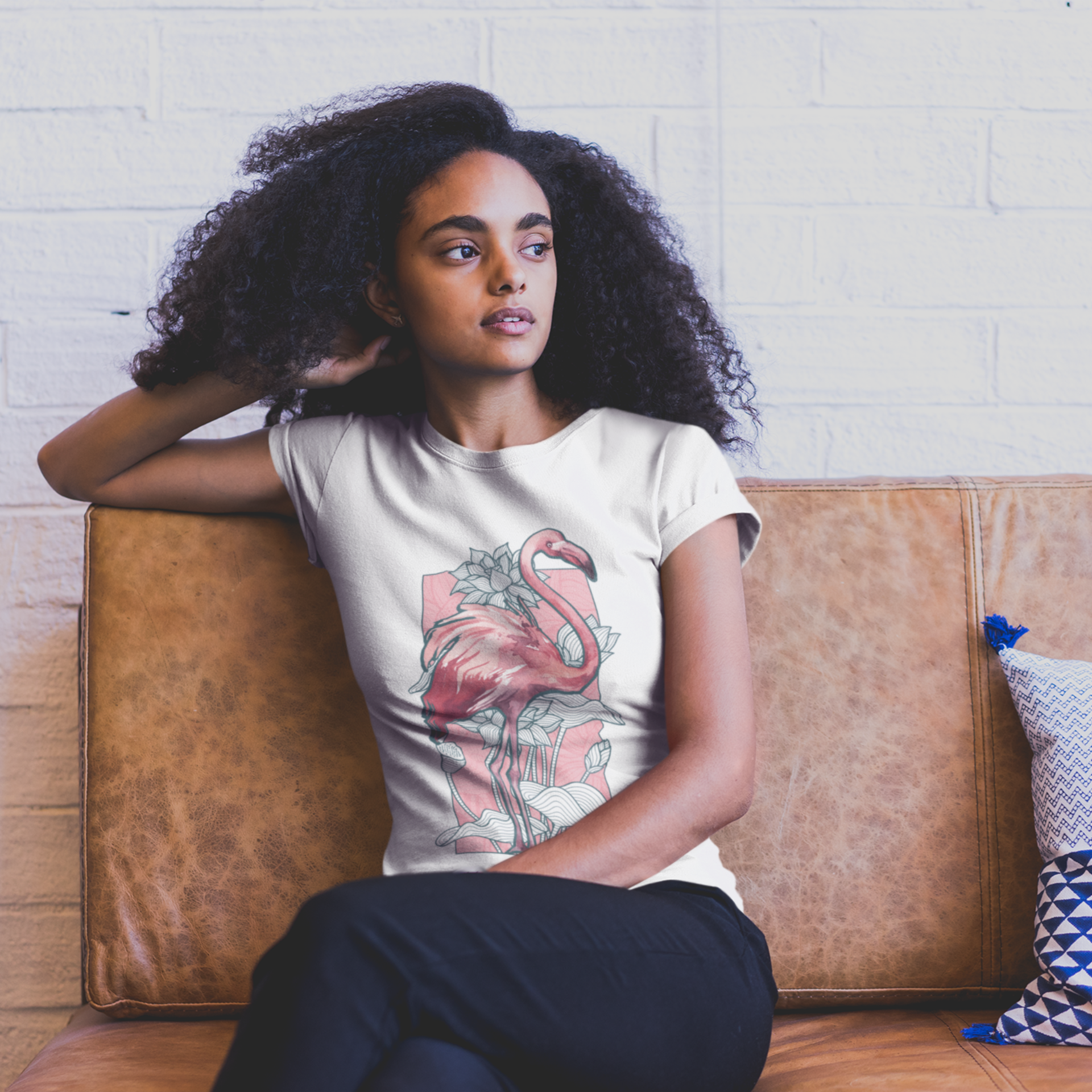 Woman sitting on a brown couch wearing a white t-shirt with a flamingo design, against a light brick wall.