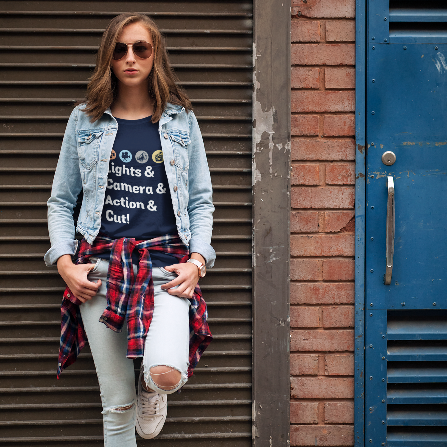 Woman standing in front of a metal shutter and blue door wearing a denim jacket, graphic t-shirt, plaid shirt, and ripped jeans.