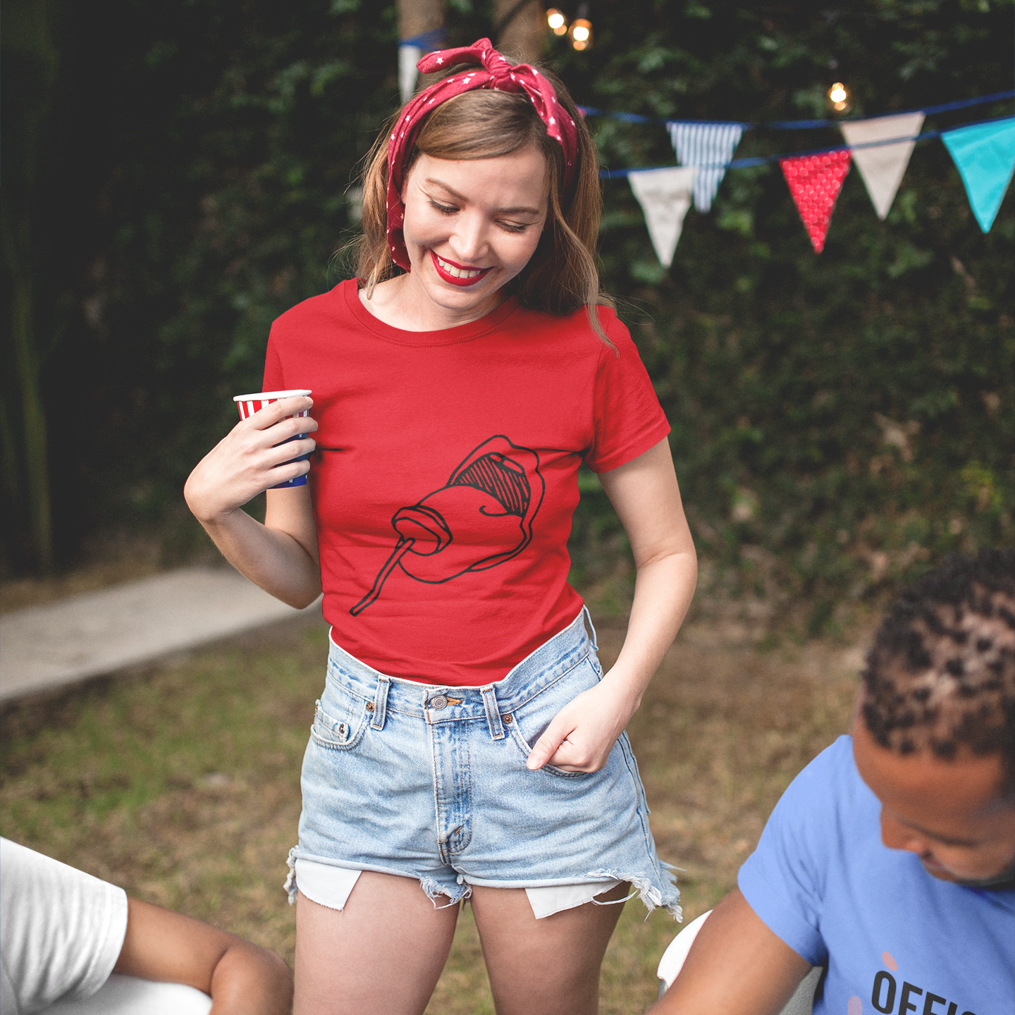 Woman in a red t-shirt with a graphic design, holding a cup outdoors with bunting flags in the background.
