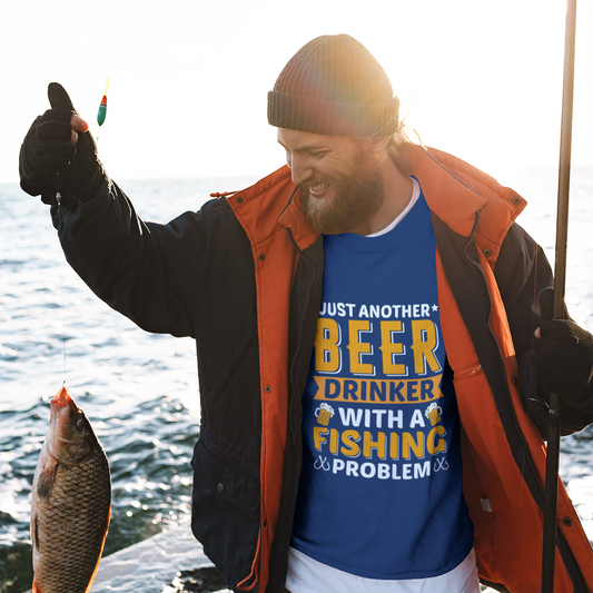 Man holding a fish and a fishing rod by the water, wearing a blue t-shirt with text.