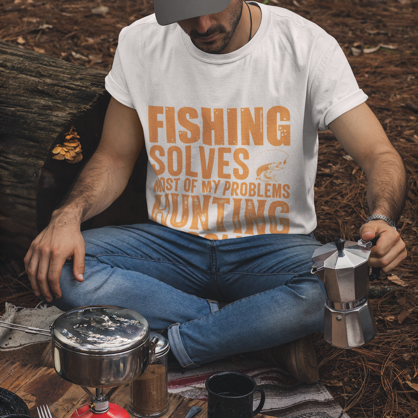 Man sitting outdoors with camping gear and a coffee maker, wearing a t-shirt with text.