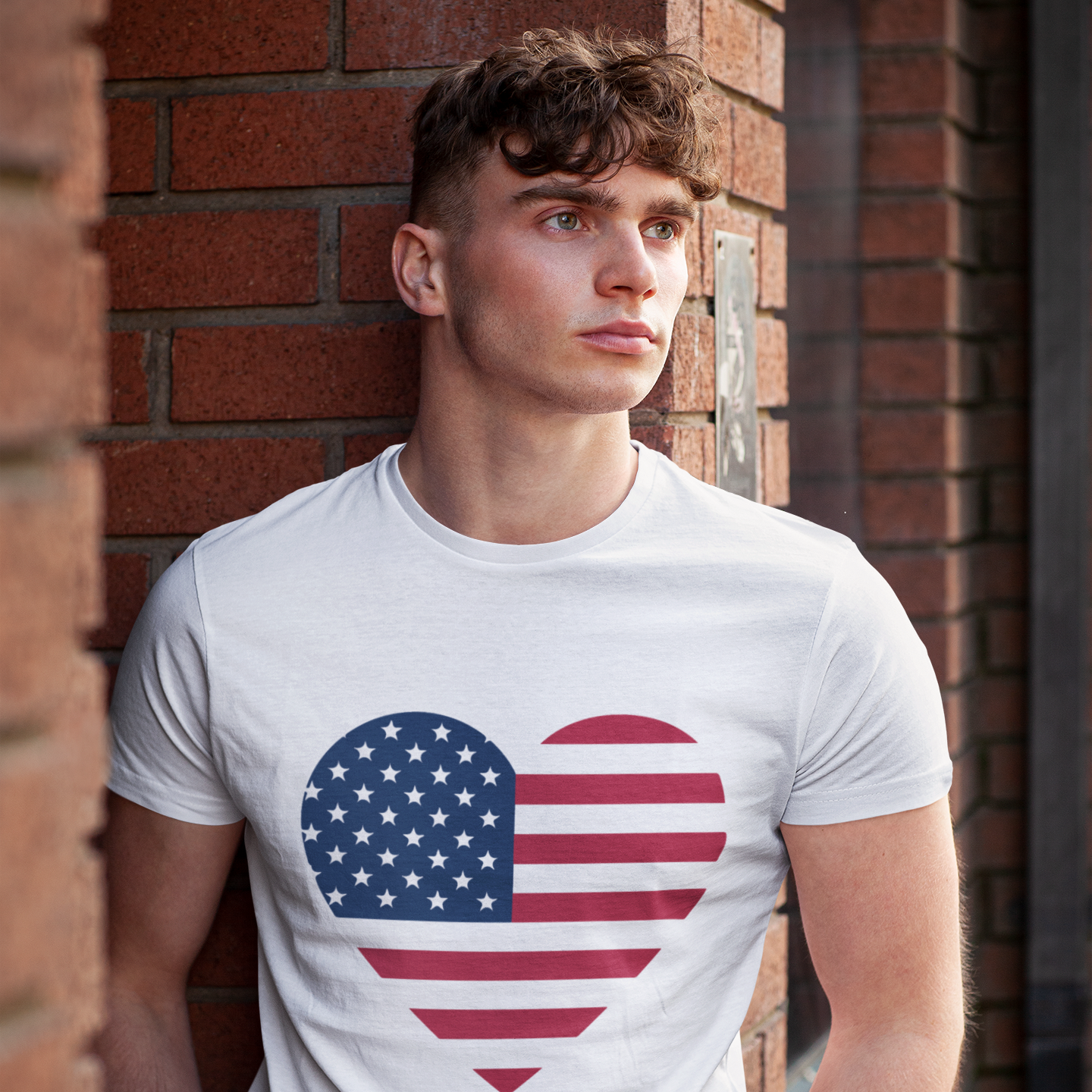 Man wearing a white t-shirt with an American flag heart design against a brick wall.