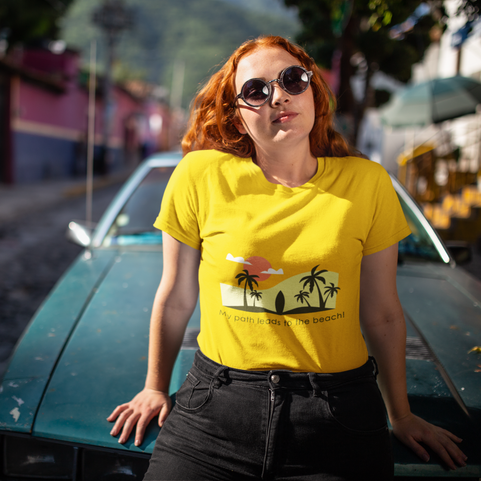 Woman wearing a yellow t-shirt with a palm tree design, standing next to a vintage car on a street.