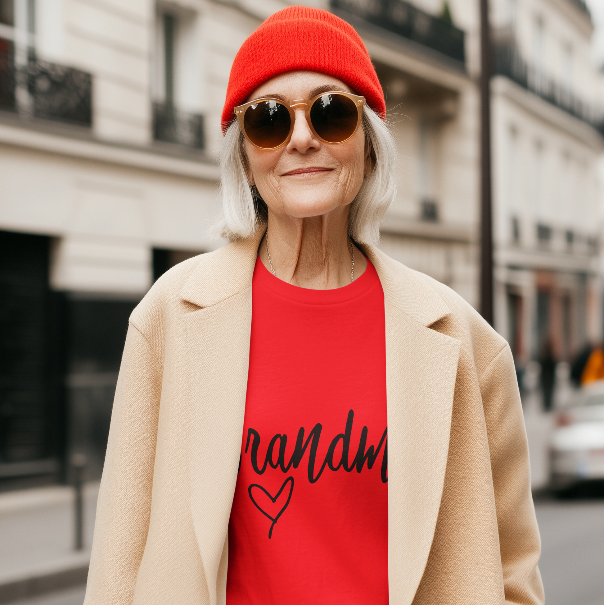 Person wearing a red beanie, sunglasses, red shirt with text, beige coat, and blue jeans on a city street.