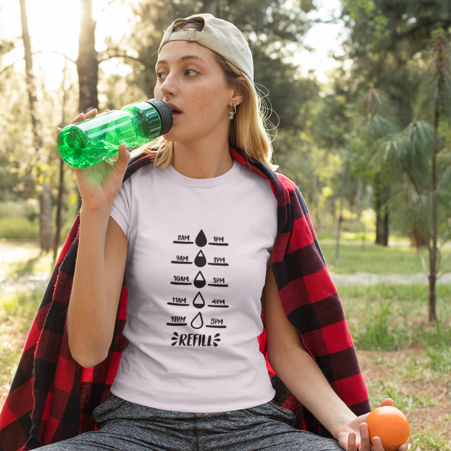 Woman sitting outdoors drinking from a green bottle, wearing a white t-shirt with a graphic design.