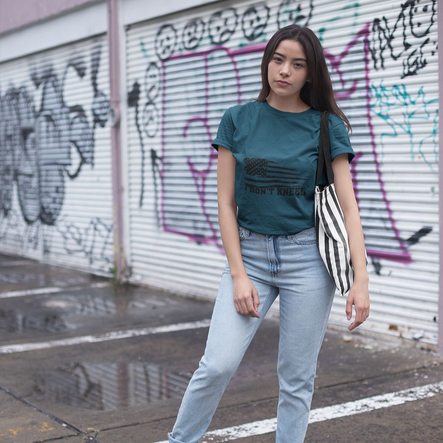 Woman standing in front of a graffiti-covered wall wearing a green t-shirt and light blue jeans.